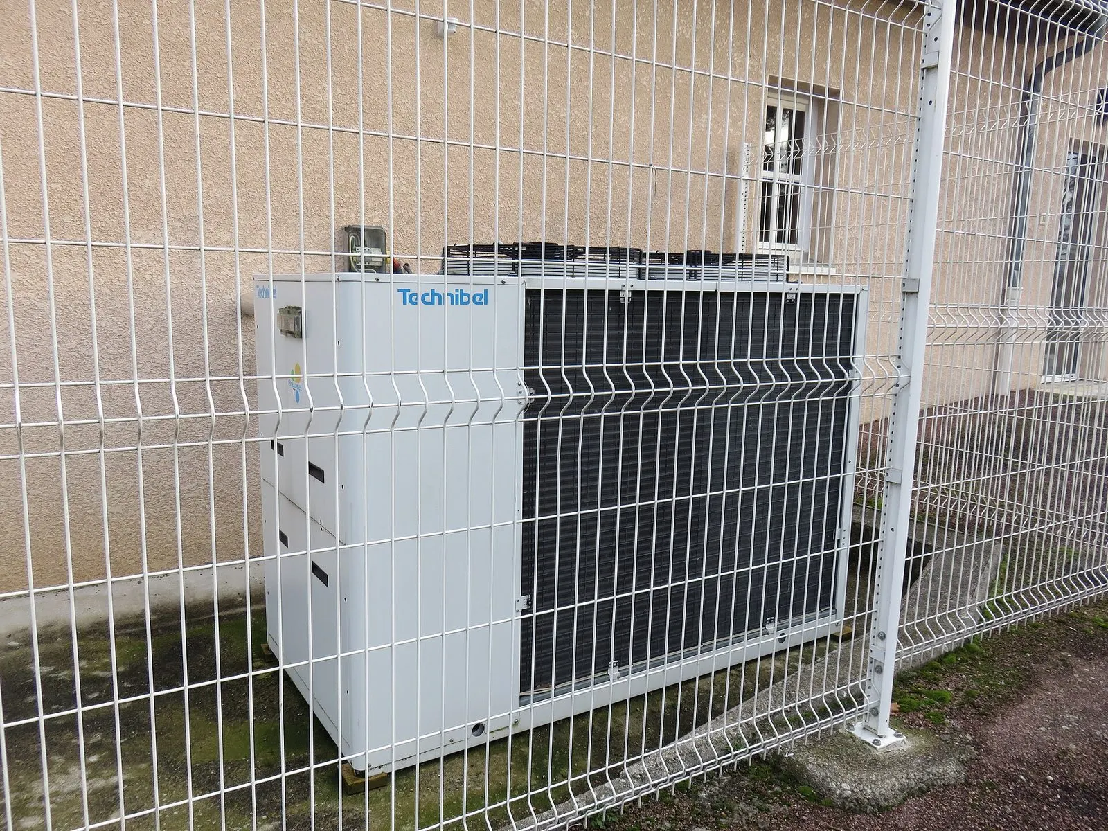 Pompe à chaleur derrière un grillage, contre un bâtiment crépi, à Saint-Vincent-de-Boisset (Loire)
